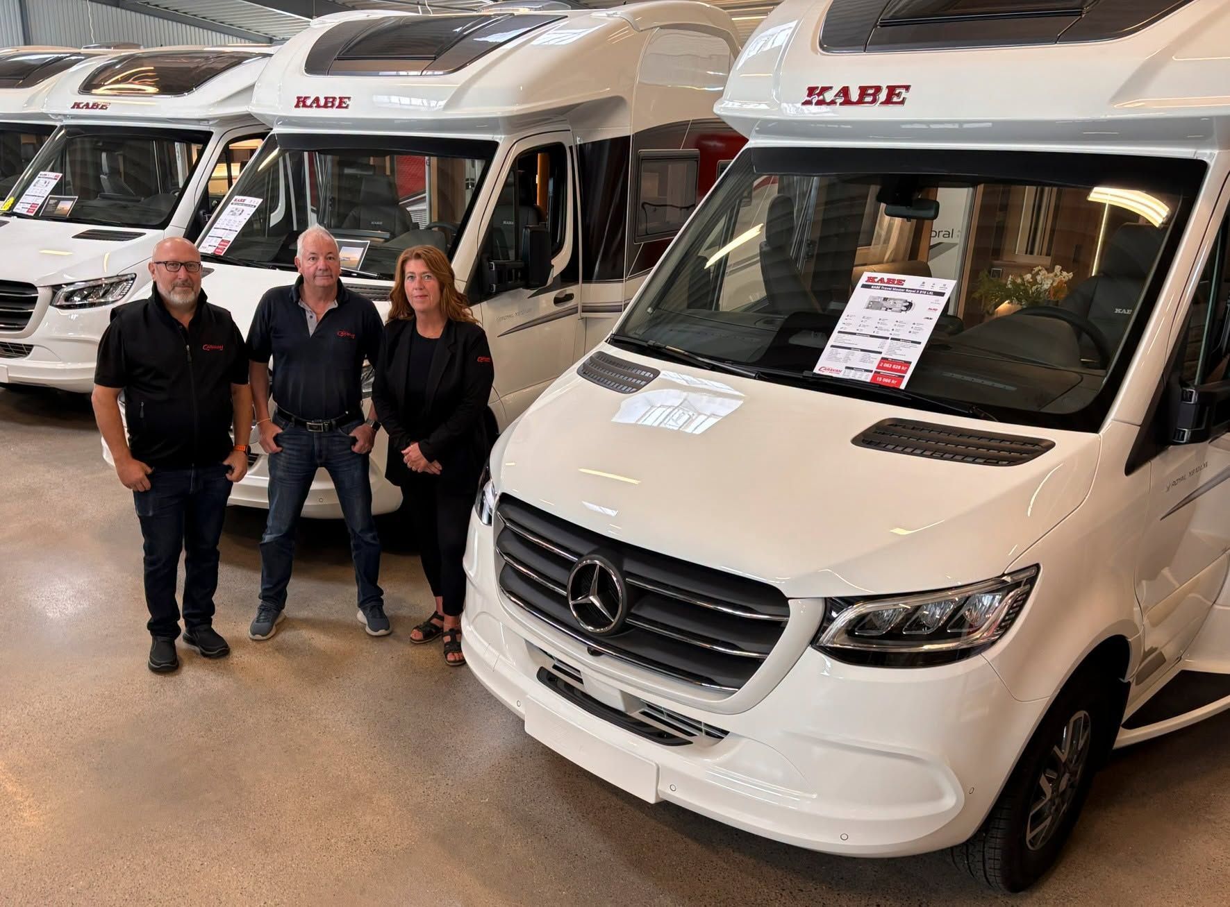 Five people stand in front of a white caravan with red accents. They are in a showroom setting.