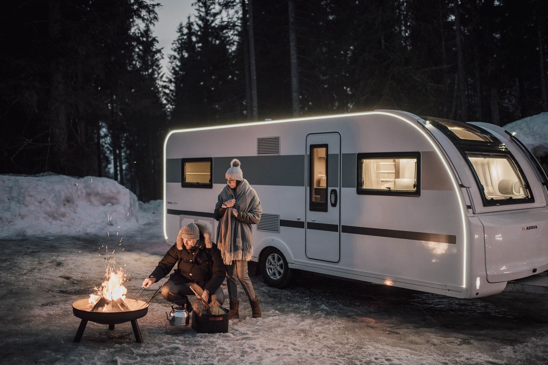 Couple camping in snowy woods with a campfire next to a lighted camper.