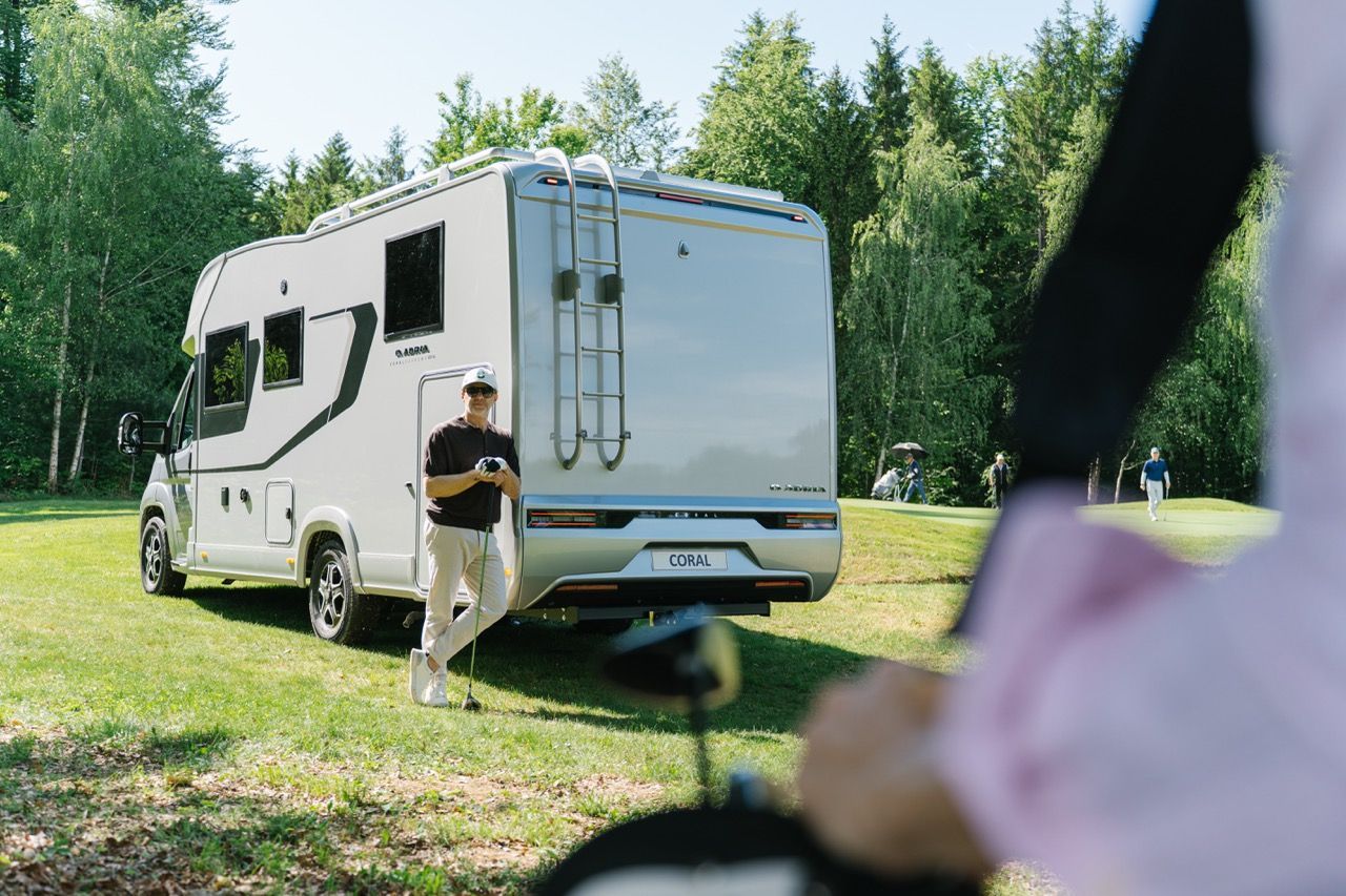 Person stands by a white camper van in a grassy park with trees in the background. Golf
