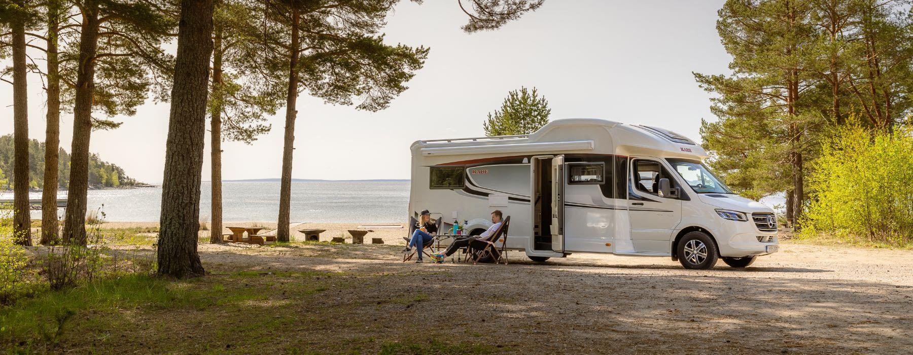 White camper van parked near a beach. Two people sit in chairs outside. Forest surrounds the scene.