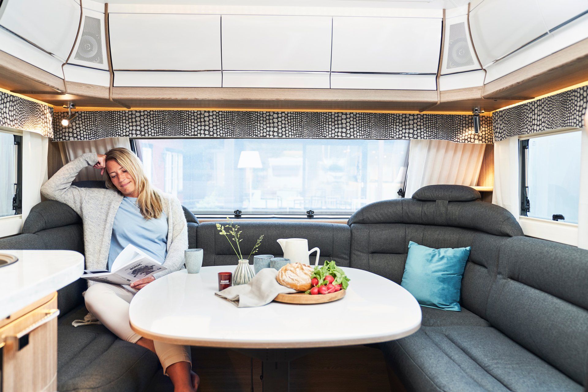 Woman relaxing in a camper interior, seated at a table with food, reading.