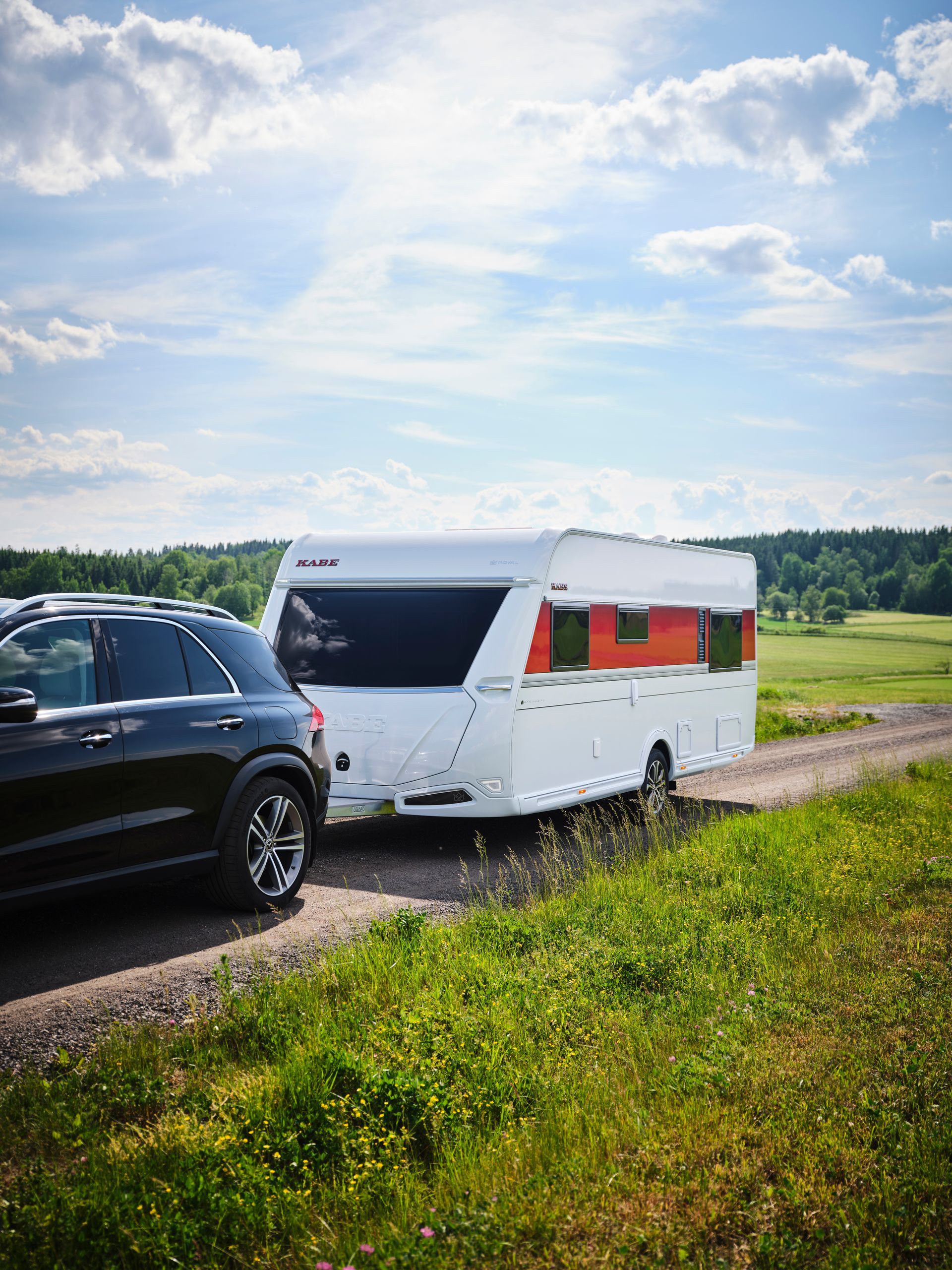 Black SUV towing a white camper with orange accents on a dirt road, sunny outdoor scene.