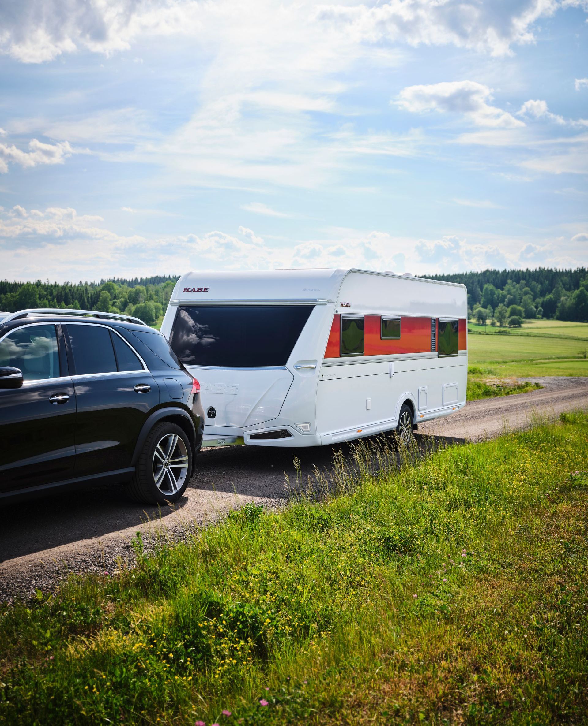 Black SUV towing a white camper on a dirt road, sunny day. Green field and trees in the background.