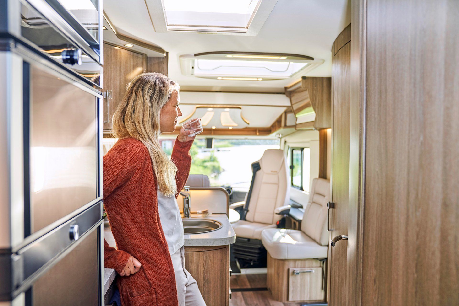 Woman in a motorhome, drinking. Interior shot with kitchen, seating, and natural light. Wooden cabinets and light upholstery.