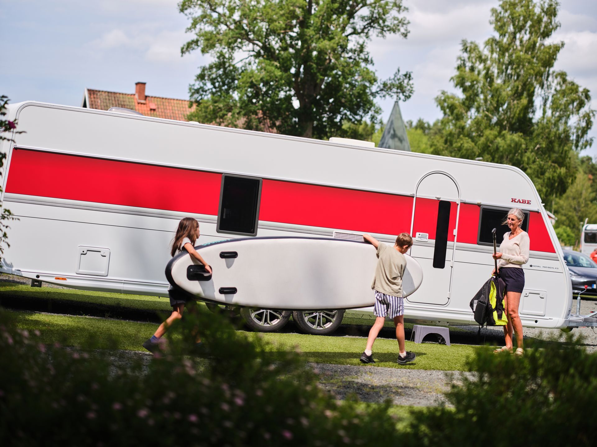 People carrying a paddleboard near a red and white caravan, in a grassy area with trees.