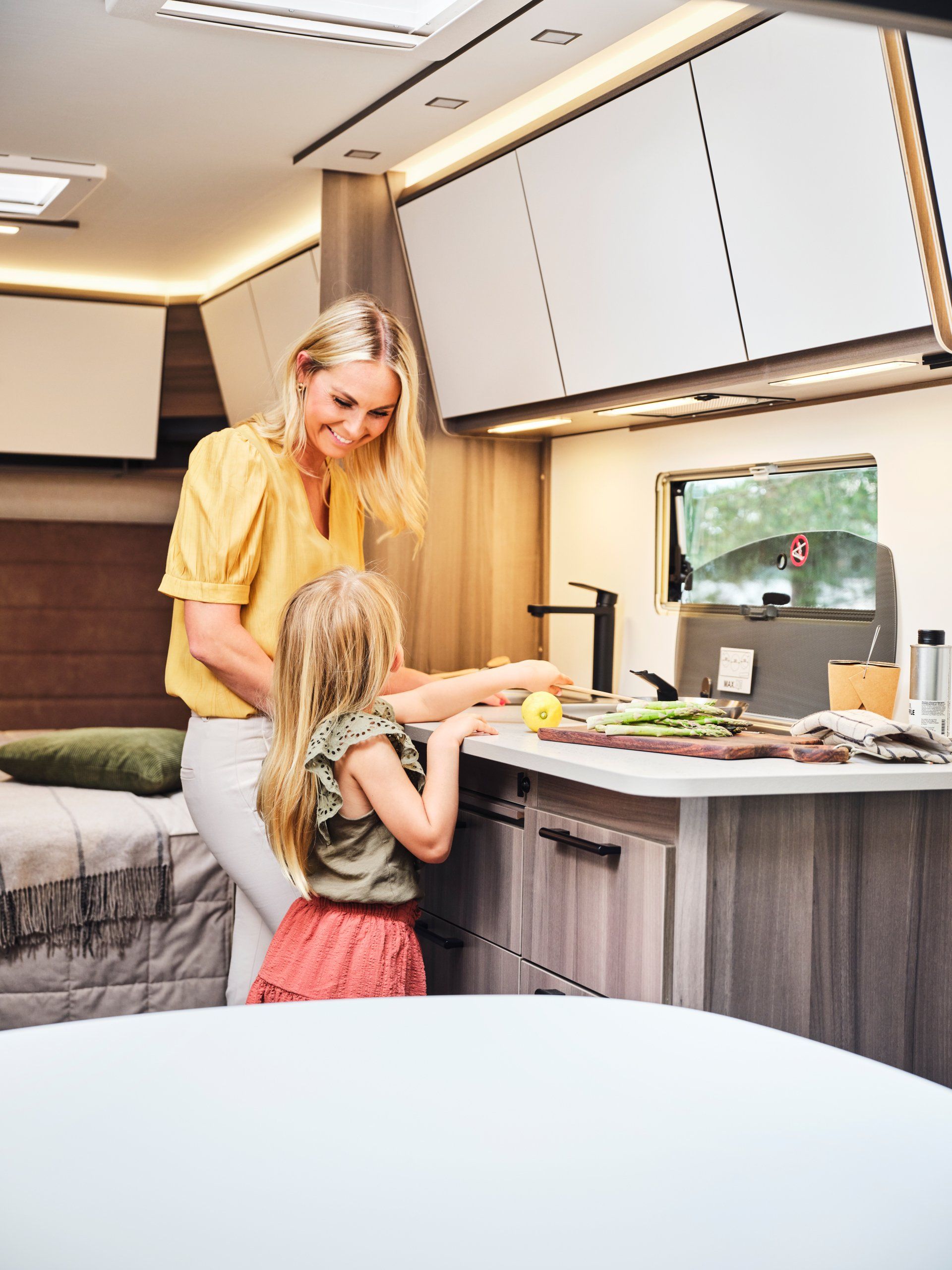 Woman and child in a camper kitchen preparing food, smiling. Interior shot, overhead cabinets, countertop.