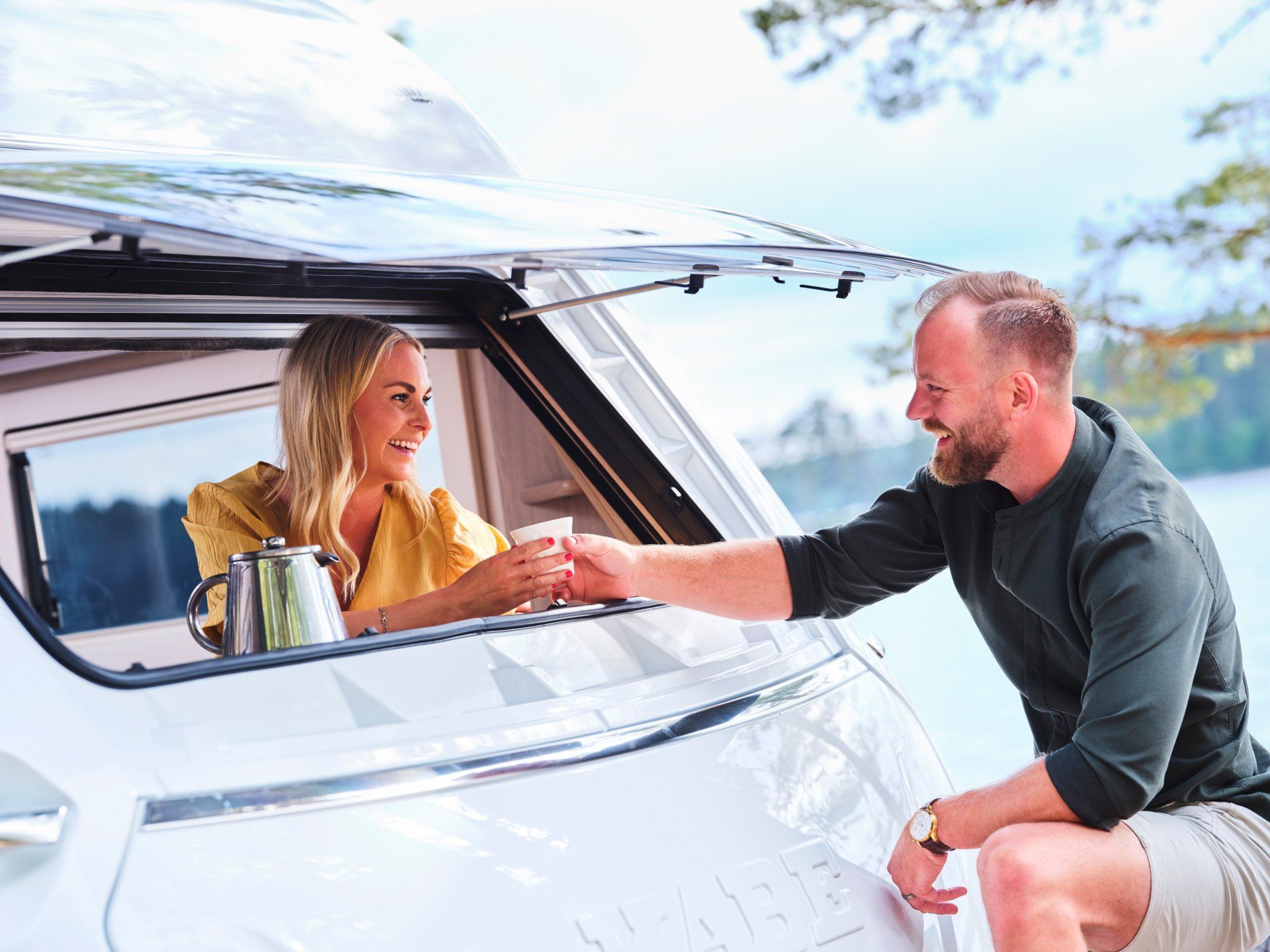 Man hands a cup to a woman in a camper van window, both smiling near a lake.