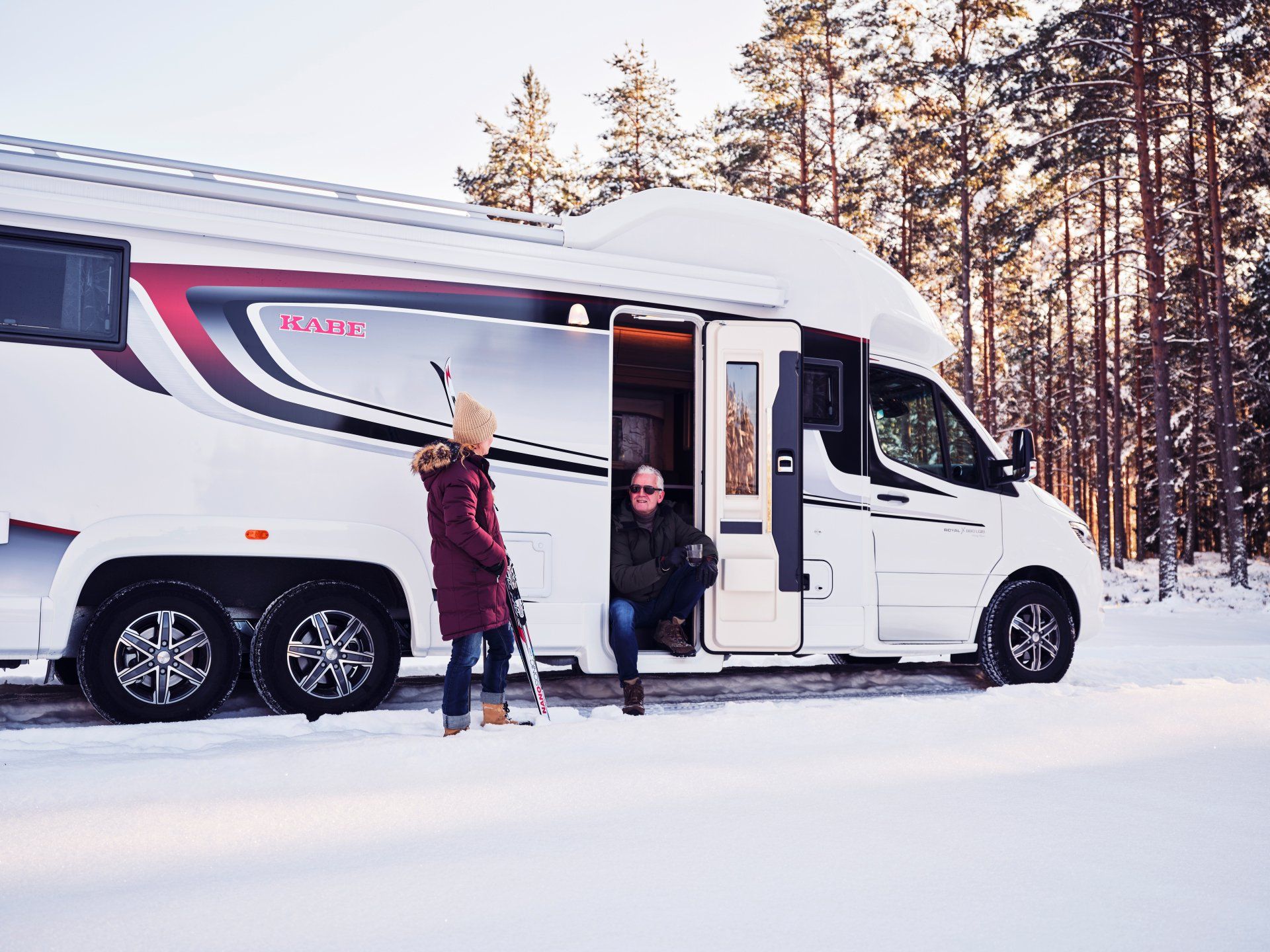 A white RV parked in snow, two people at the door: one with skis, one seated inside, trees in background.