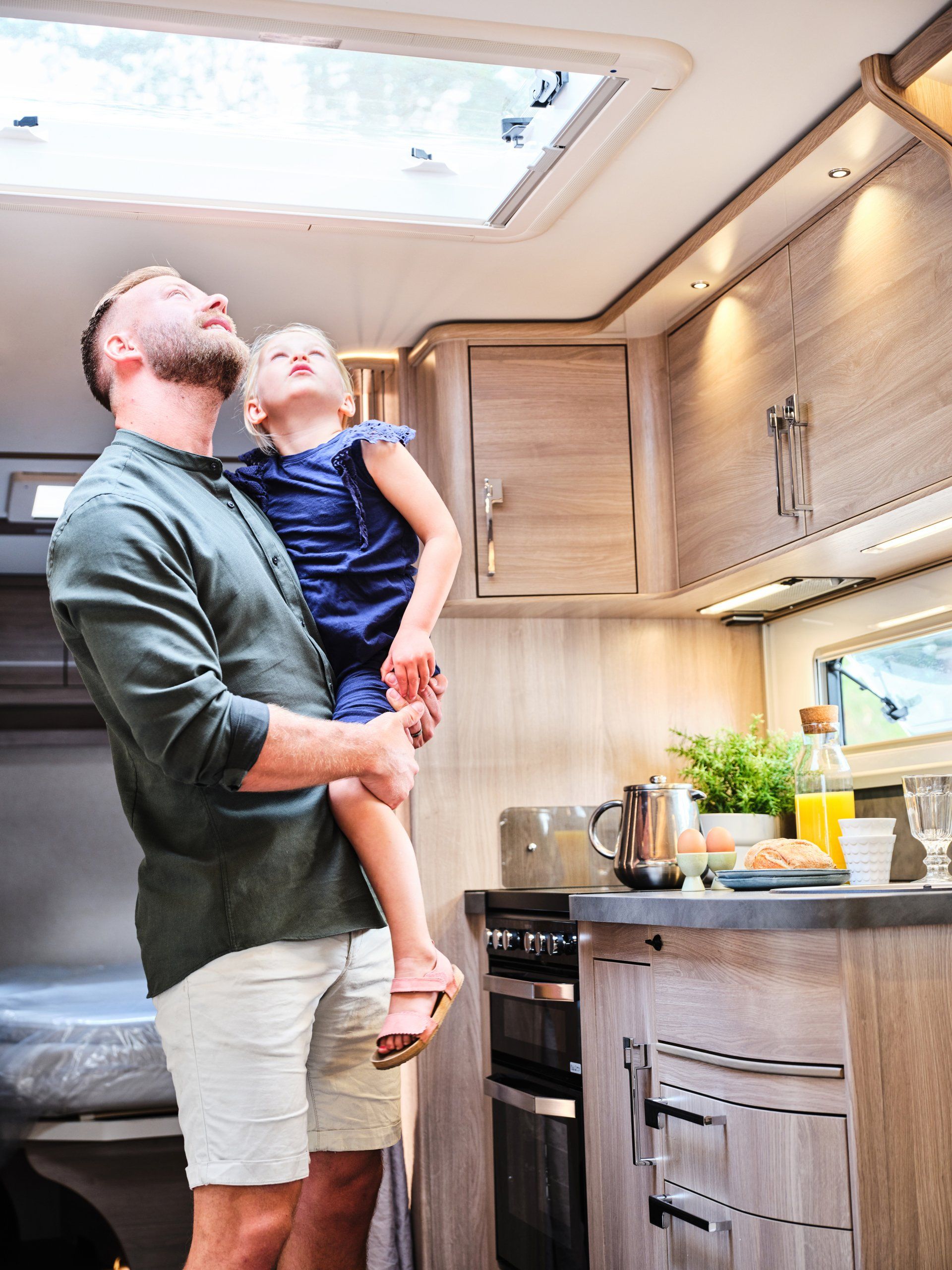 Man holding child, looking up at skylight inside a camper's kitchen; light wood cabinets, small appliances visible.
