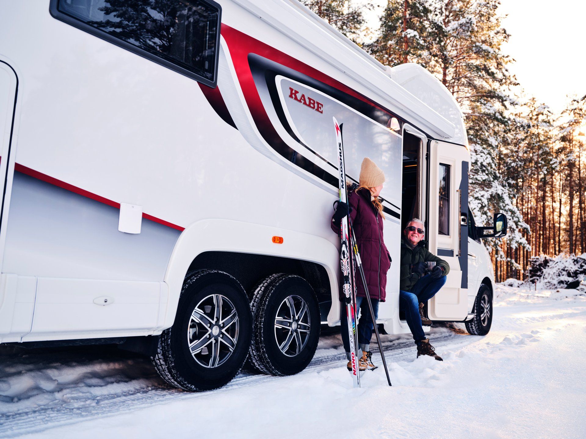 White RV with skis next to it in snow. Person outside holding skis, another in door. Red trim on the RV.