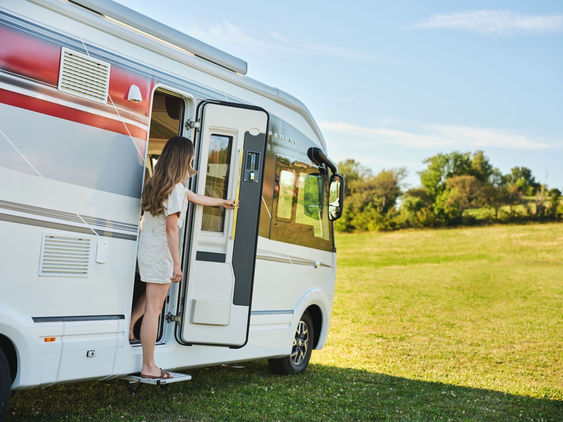 Woman steps out of a white and red RV into a grassy field on a sunny day.