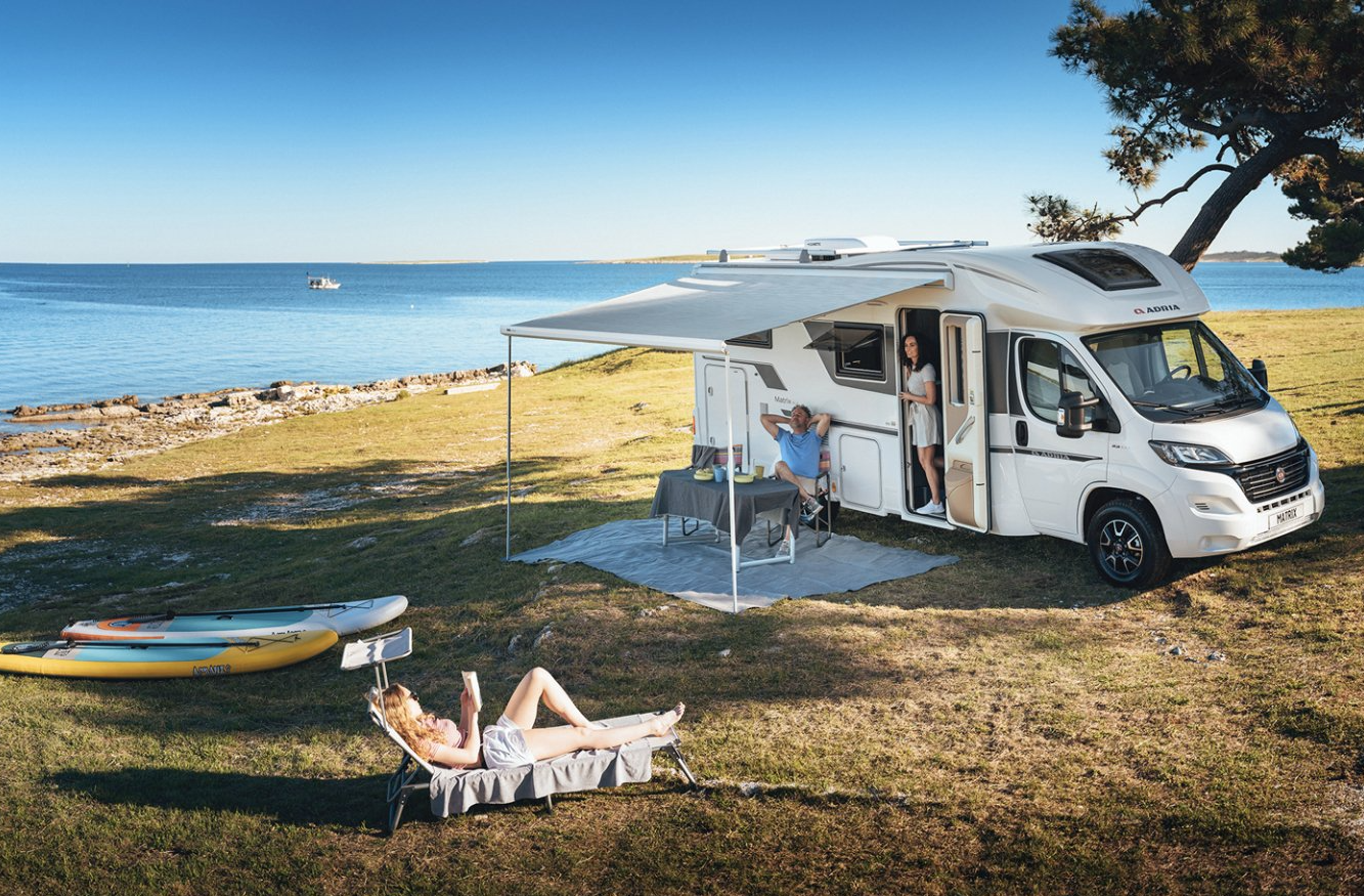 RV parked by the sea with awning extended. People relaxing; one sunbathing, others at a table. Kayaks on grass.