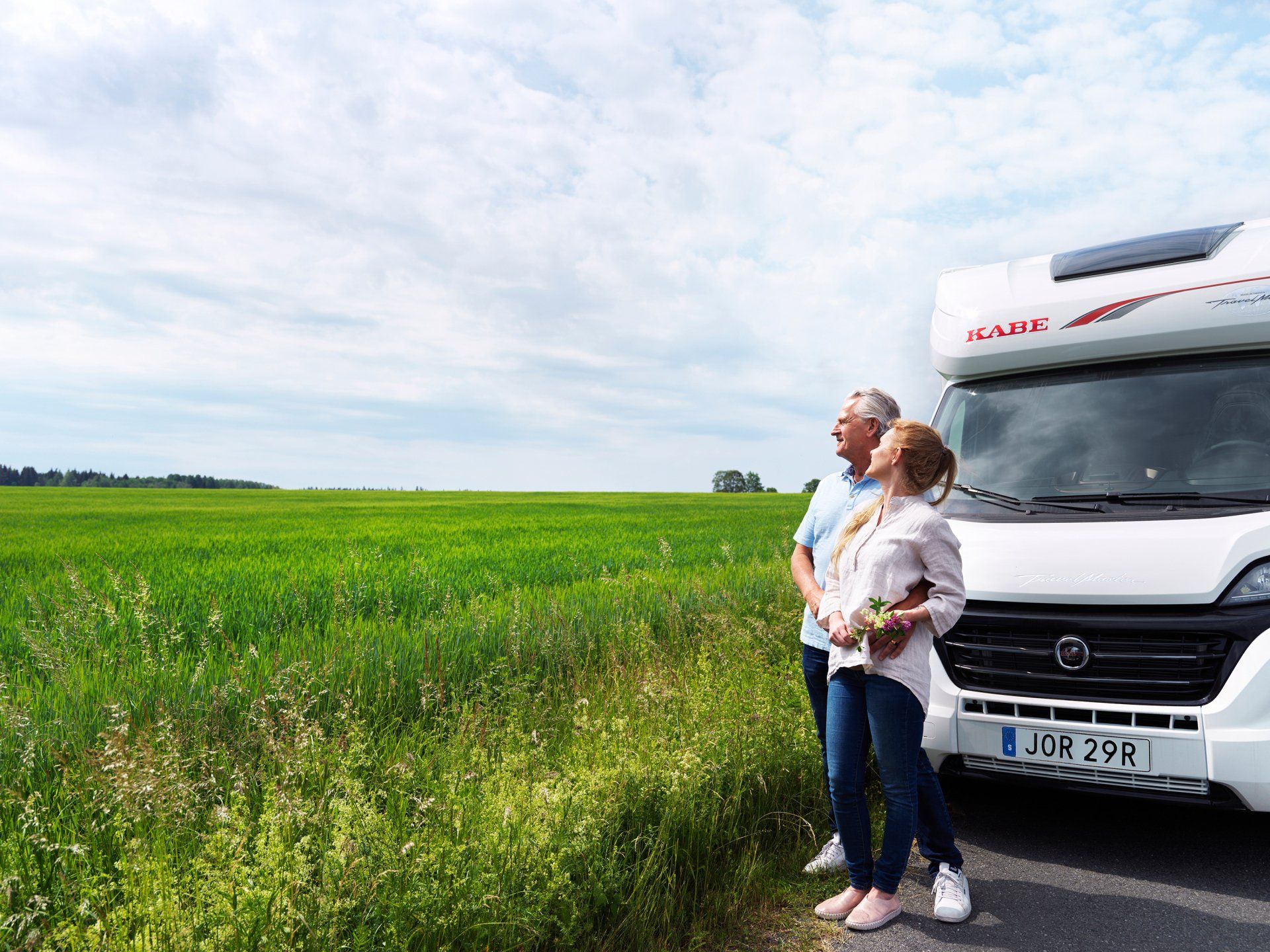 Couple standing by RV, looking at a green field under a blue sky.