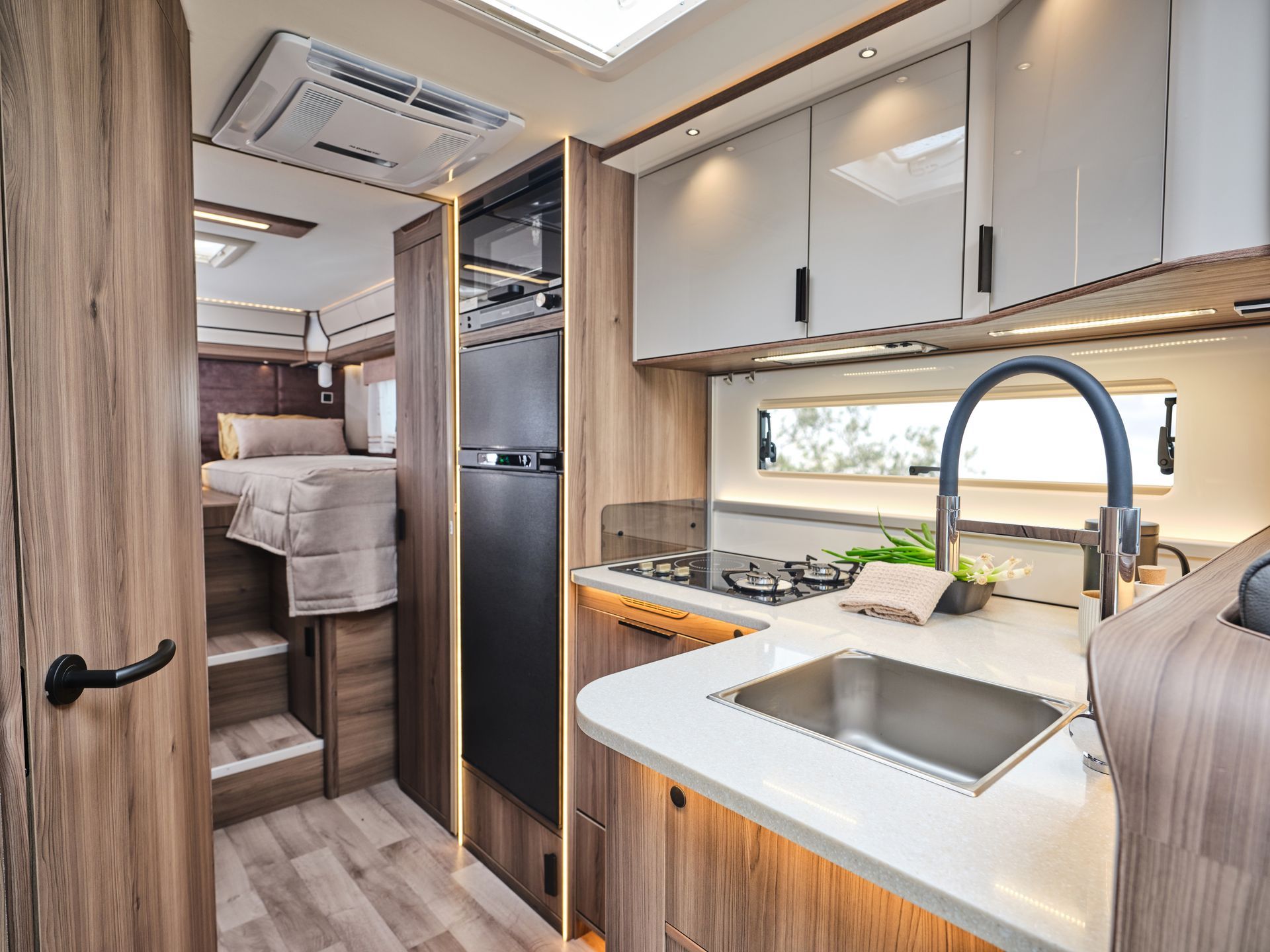 Interior of a camper, kitchen area with sink, stove, refrigerator, cabinets, and a bed in the background.