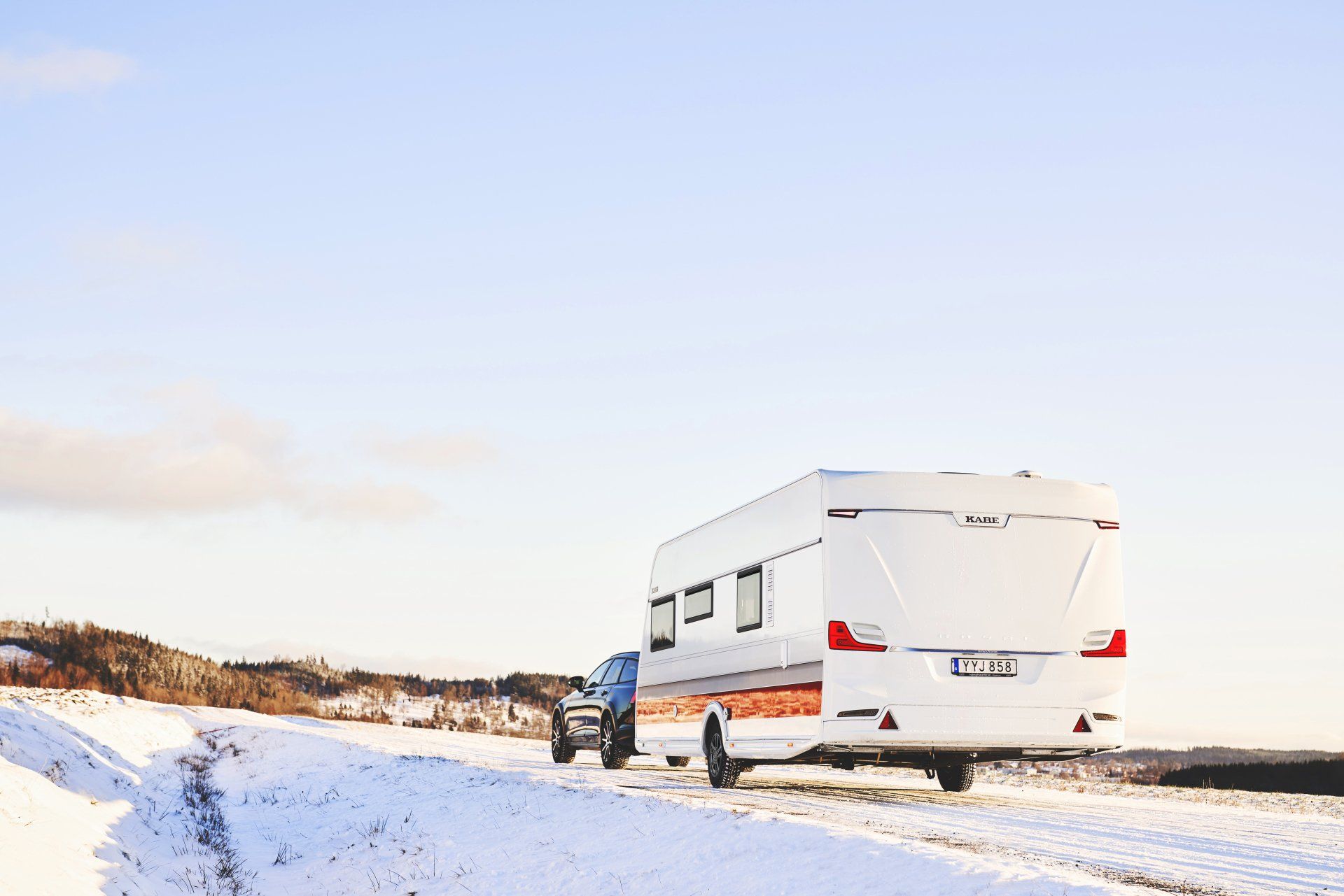 A white camper trailer being towed by a vehicle on a snowy road, sunny day.