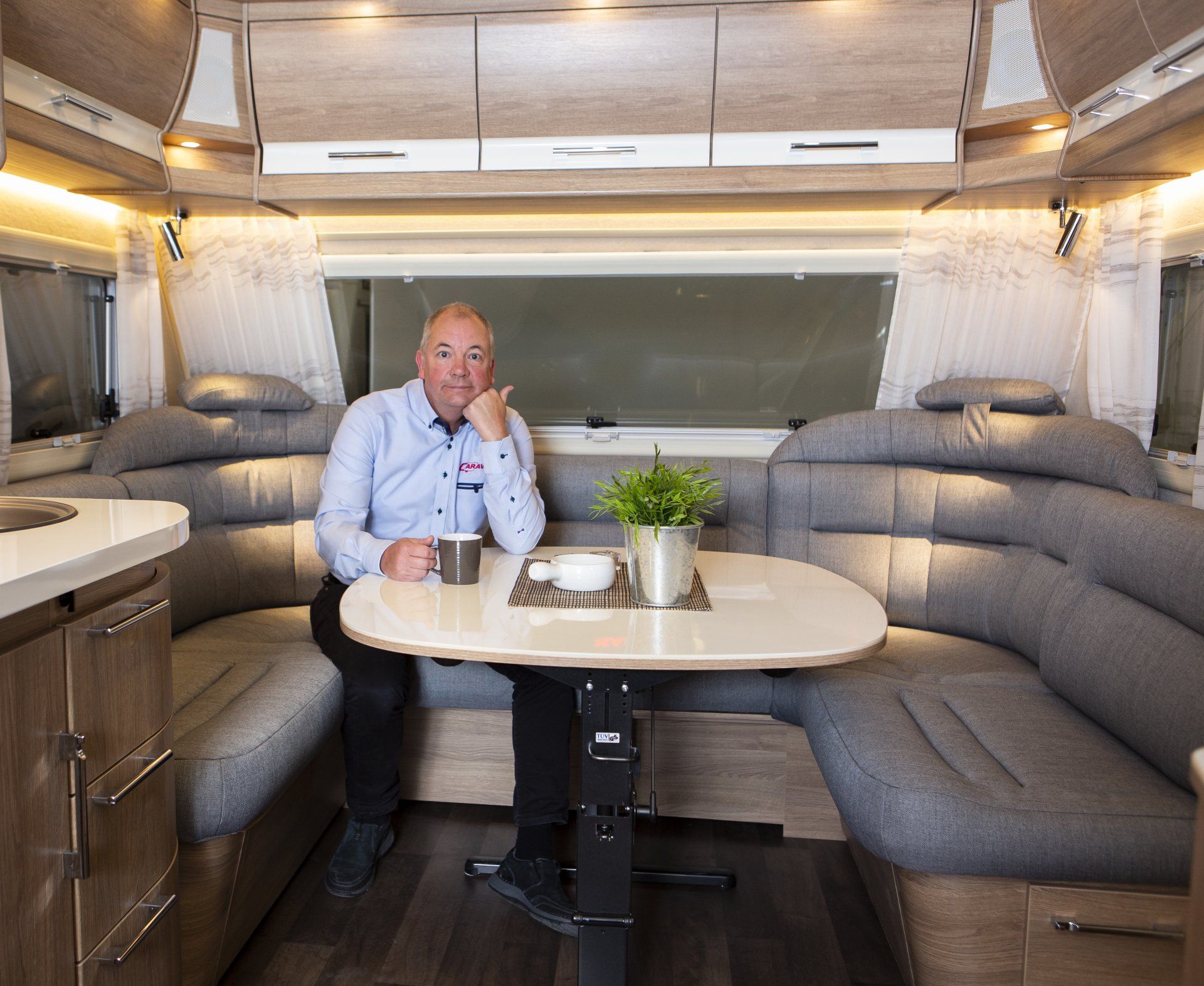 Man seated at a table inside a camper. He's holding a mug. Interior has grey seating, wooden cabinets, and a large window.