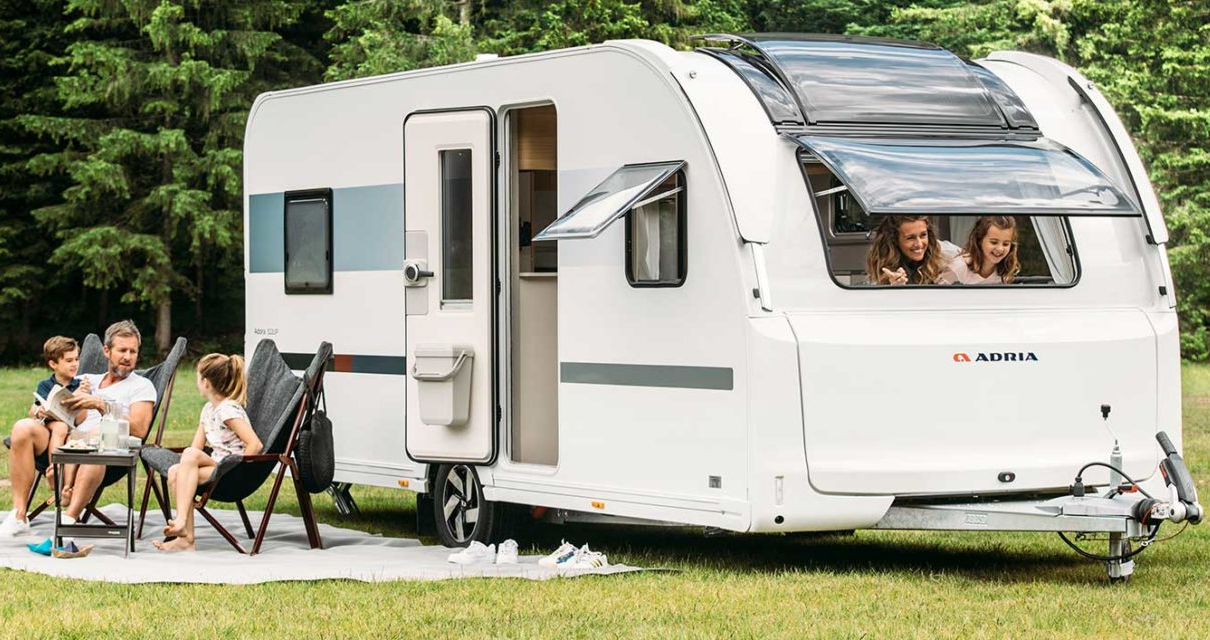 White travel trailer with a family sitting outside; green grass, open door, clear windows.