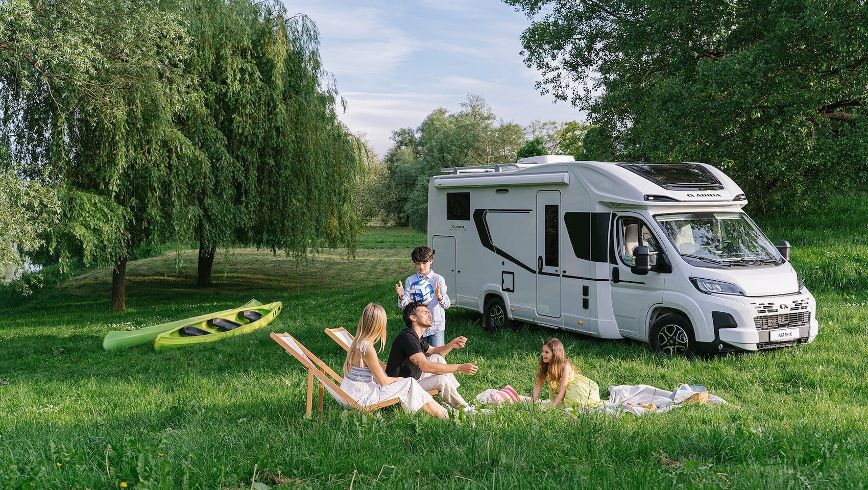 Family relaxing on blankets beside a white camper van in a sunny green meadow
