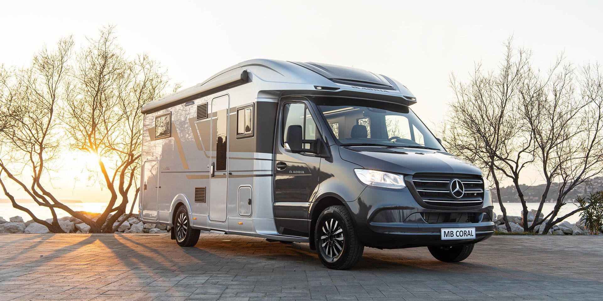 Gray and white camper van parked on a rocky shoreline with trees and water in the background at sunset.