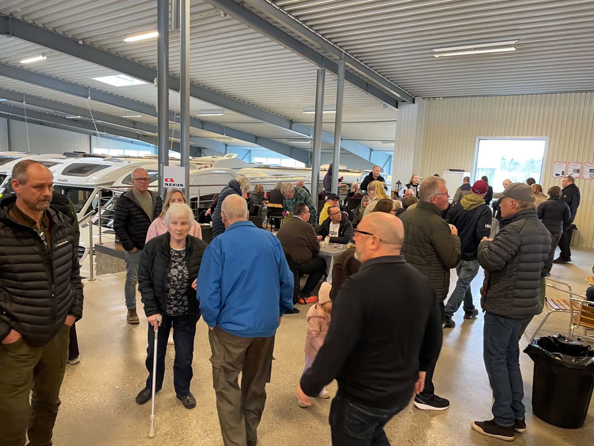 People gathered in a bright indoor hall, talking and standing near metal pillars and a crowd in the background