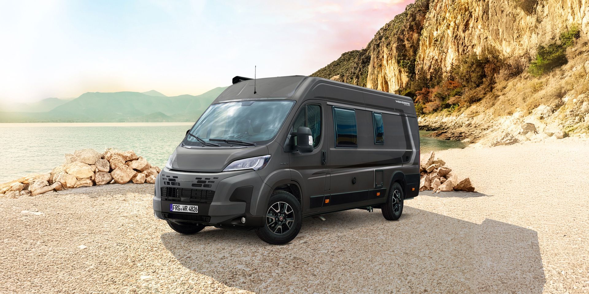 A dark gray camper van on a sandy beach, with a mountain in the background.