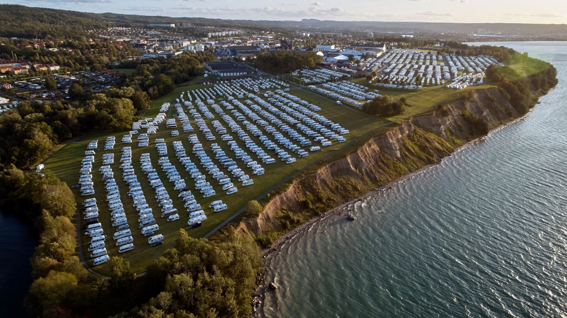 Aerial view of RVs and campers densely packed along a lakeside cliff, with a town visible in the distance.