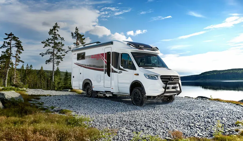 White campervan on gravel path near a lake and trees.