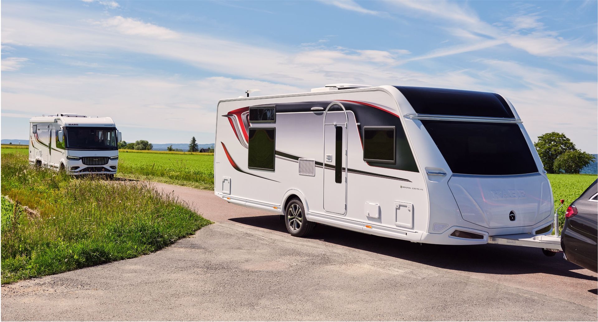 White travel trailer parked on a road beside a field; another RV is visible in the distance.