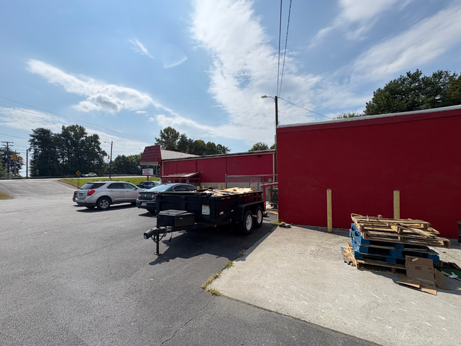 Outdoor parking lot beside a red building with parked cars, trailers, and stacks of pallets under a partly cloudy sky