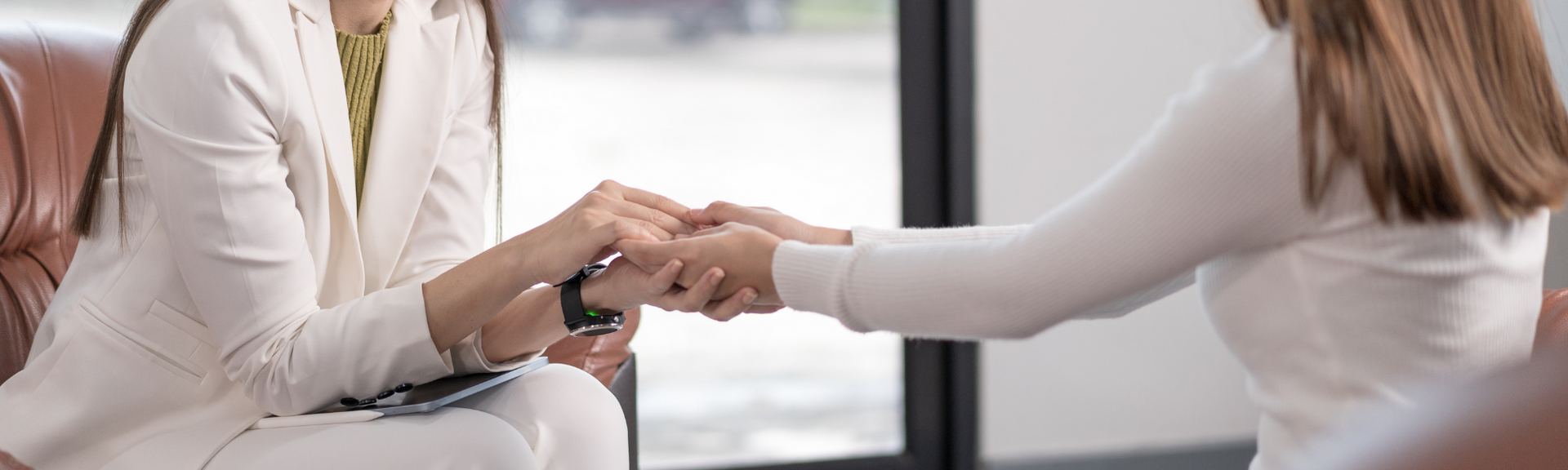 Two people sit in a room holding hands in a supportive gesture, focusing on their joined hands.