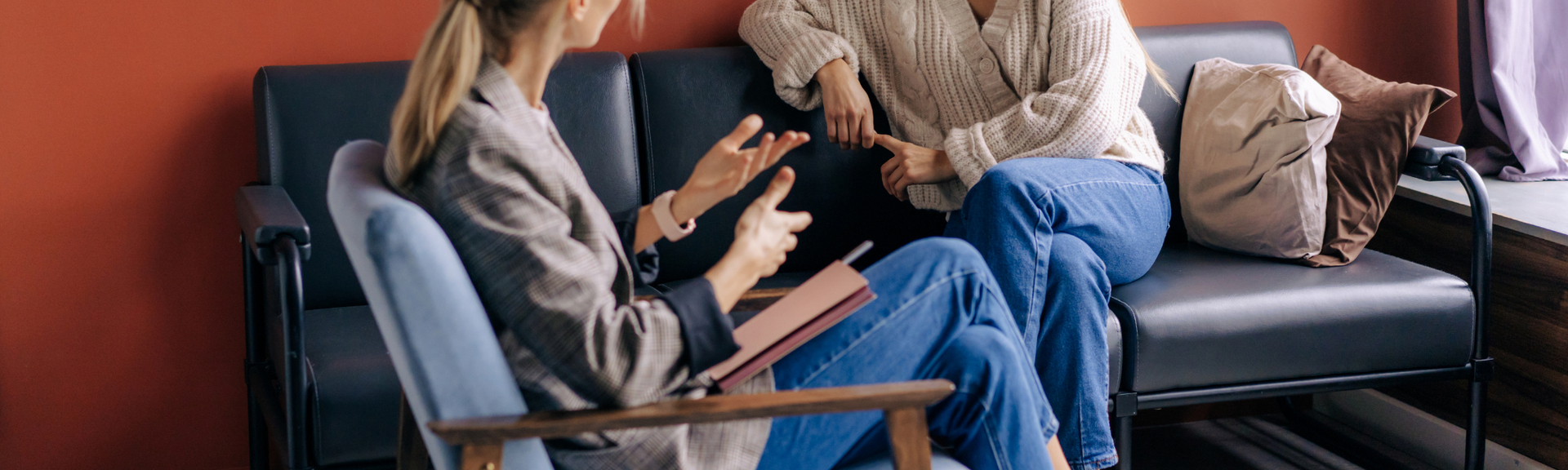 A person in a blazer sits in a chair, gesturing while talking to someone on a sofa in a room with reddish walls.