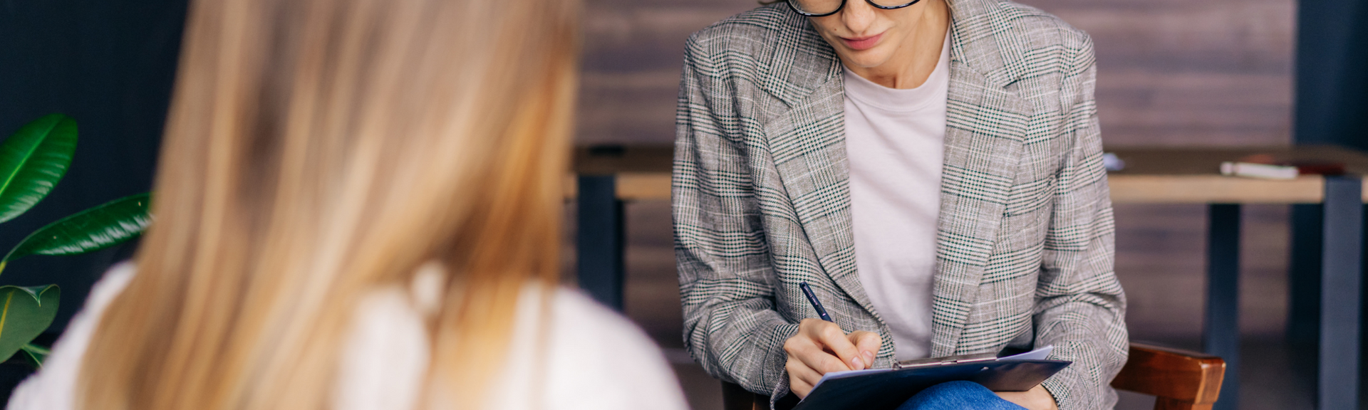 A person in a patterned blazer sits across from another individual, writing on a clipboard during a consultation.