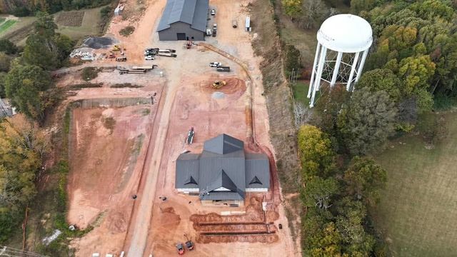 Aerial view of a house under construction beside a dirt road and a white water tower, surrounded by trees.