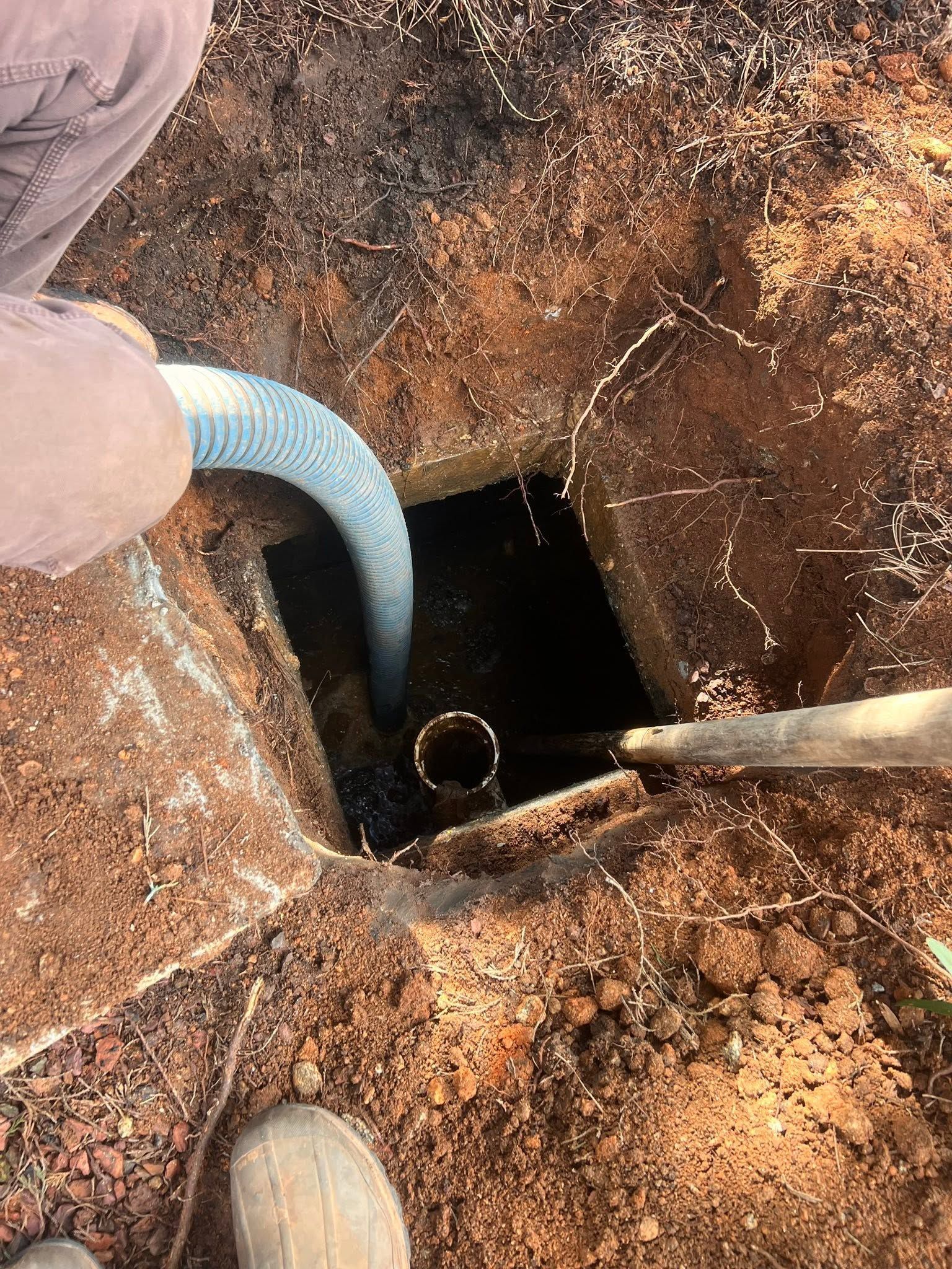 Person lowering a blue hose into a dark square well opening in a rusty ground surface