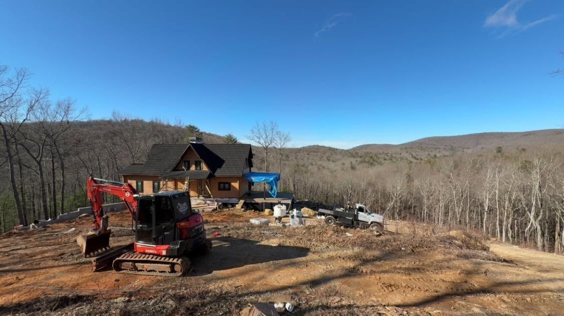 Red excavator and blue truck at a rustic cabin construction site on a hillside with wooded mountains beyond