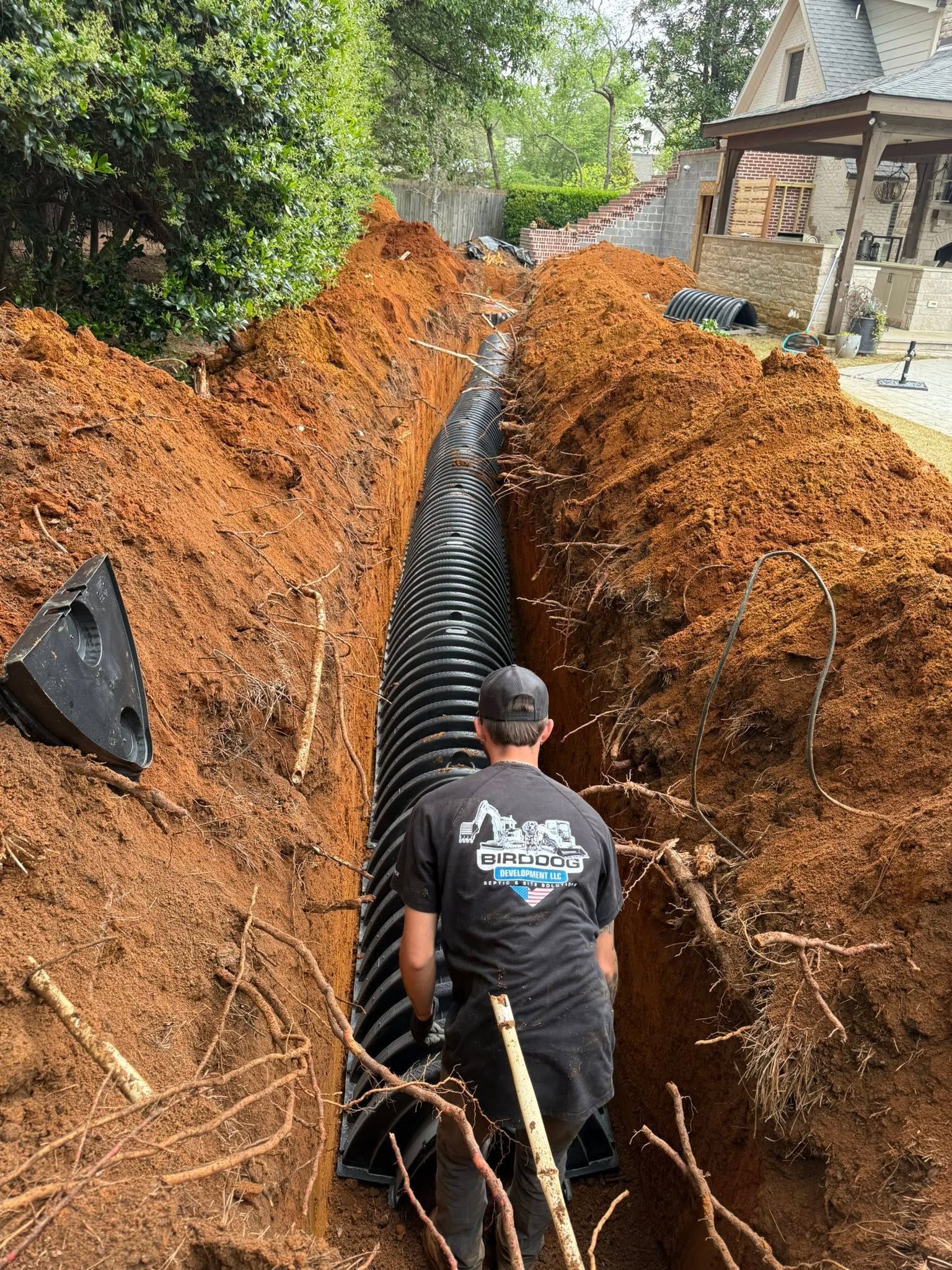 Worker installing a large black drainage pipe in a deep trench beside a house, with orange dirt walls.