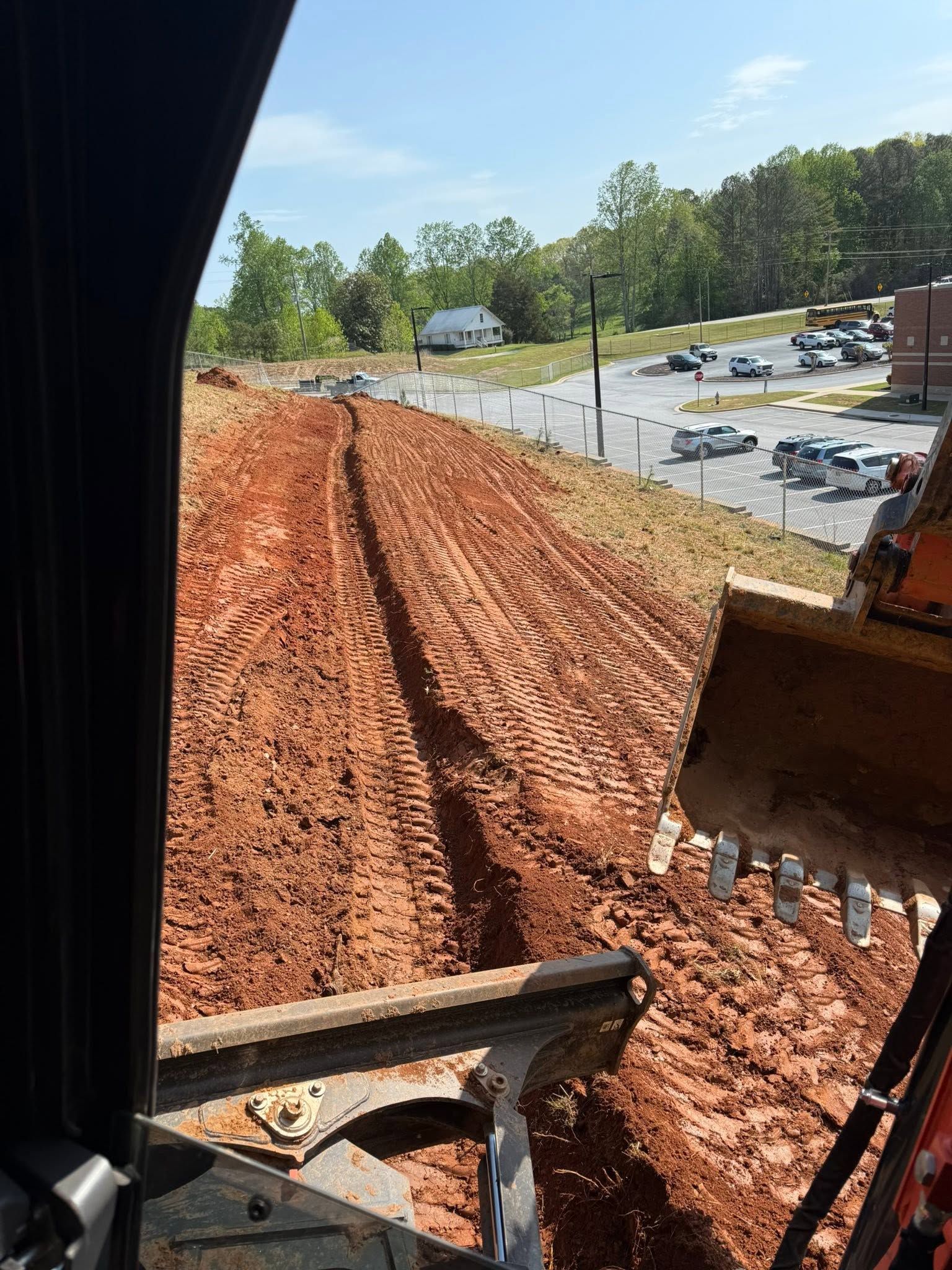 Construction site with a red dirt embankment, excavator bucket, and parking area in the background