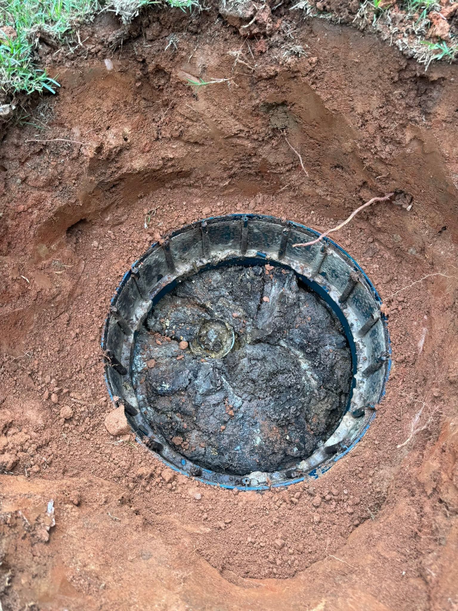 Round metal cover set in reddish-brown dirt trench, viewed from above