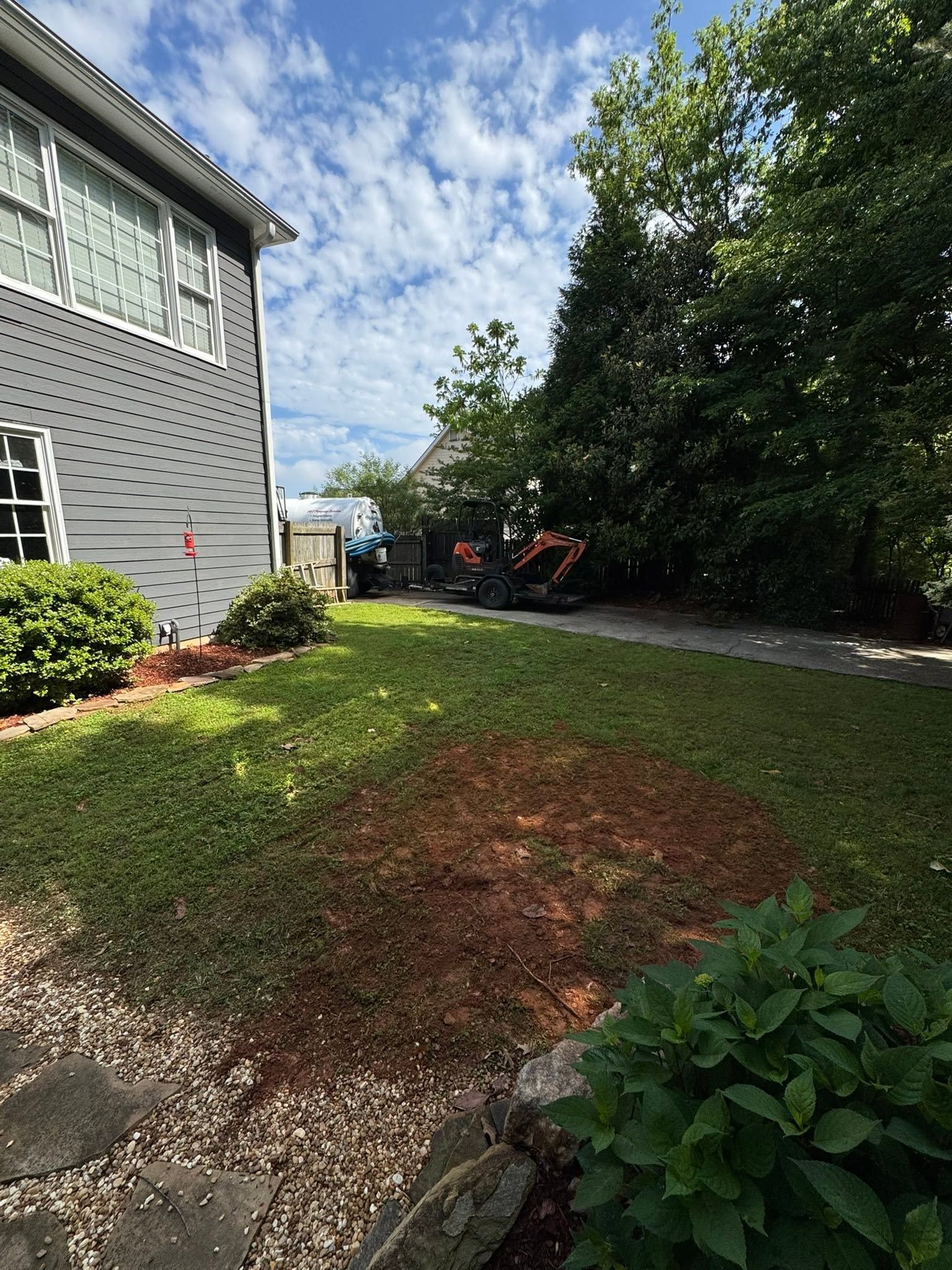 House side yard with green grass, gravel path, shrubs, and trees under a partly cloudy sky