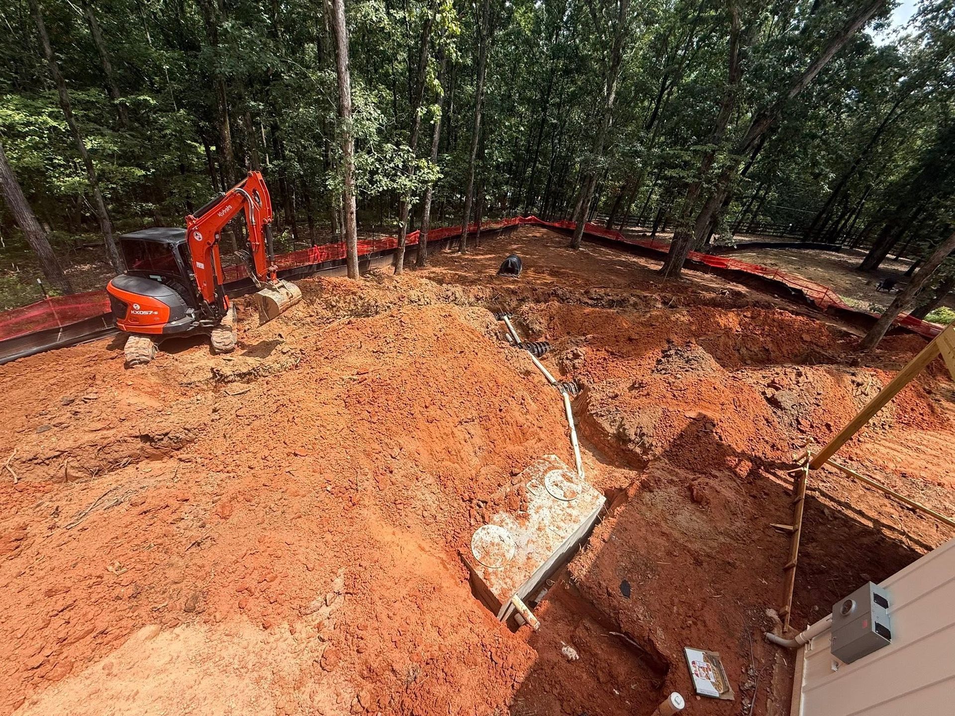 Excavator digging red soil at a wooded construction site with a large excavation pit