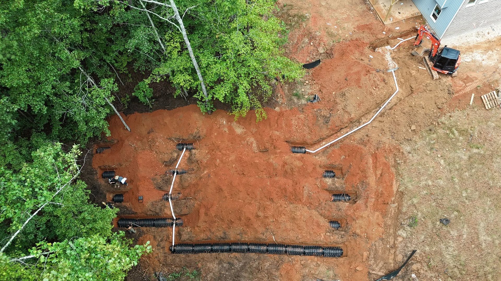 Aerial view of a dug-up red dirt area beside trees, with pipes and a small excavator near the top right.