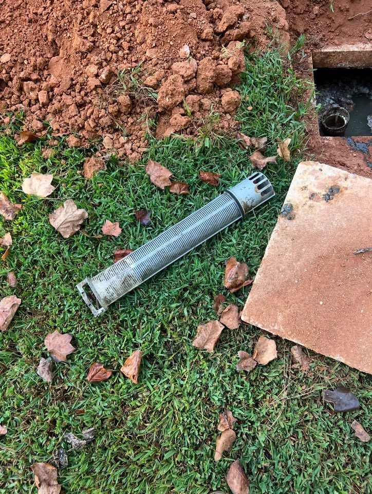 Gray pipe lying on grass beside a dirt pile, broken concrete, and an open drain hole.