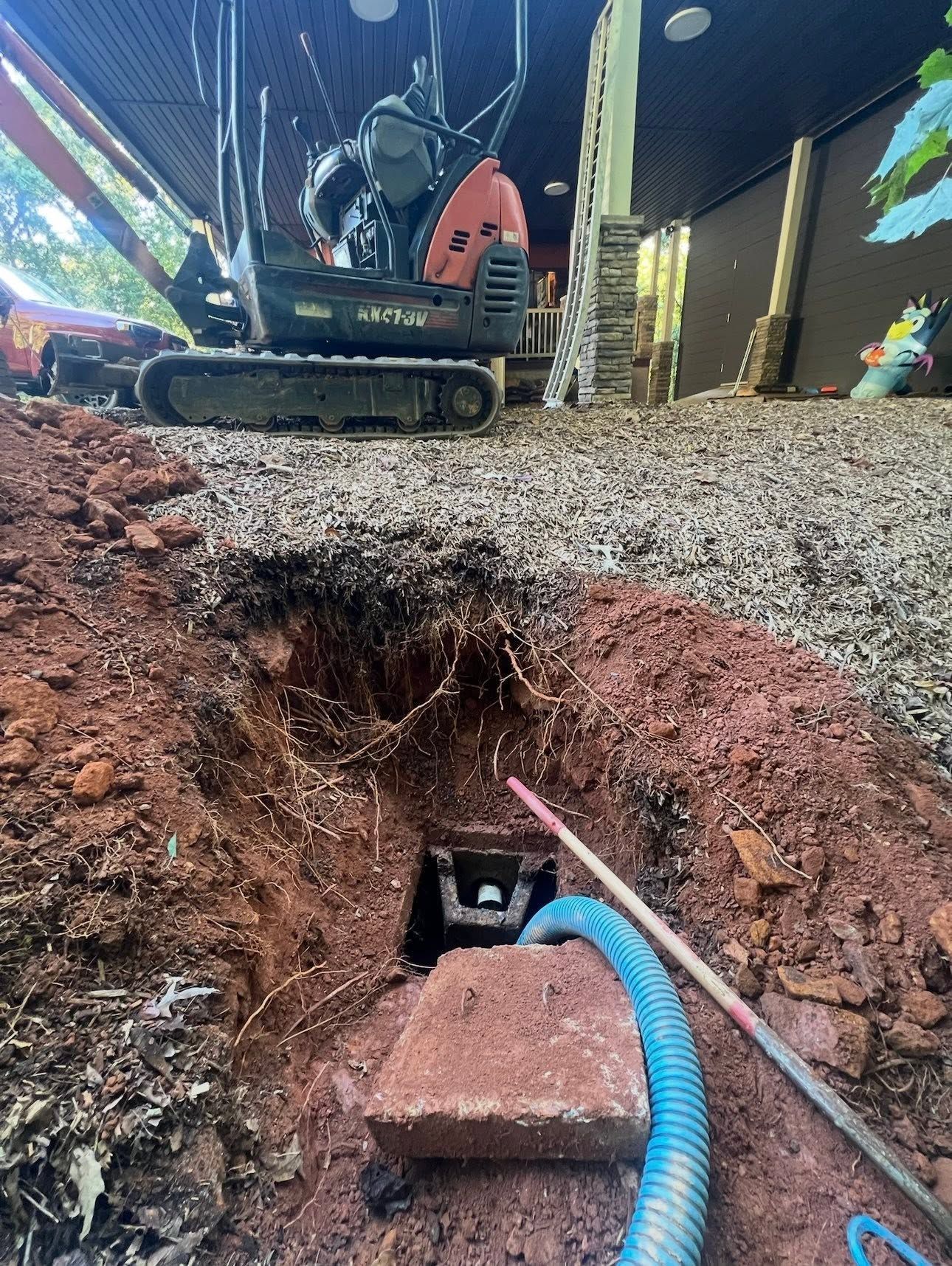 Excavator digging a trench beside a gravel surface, with a blue corrugated pipe exposed underground.