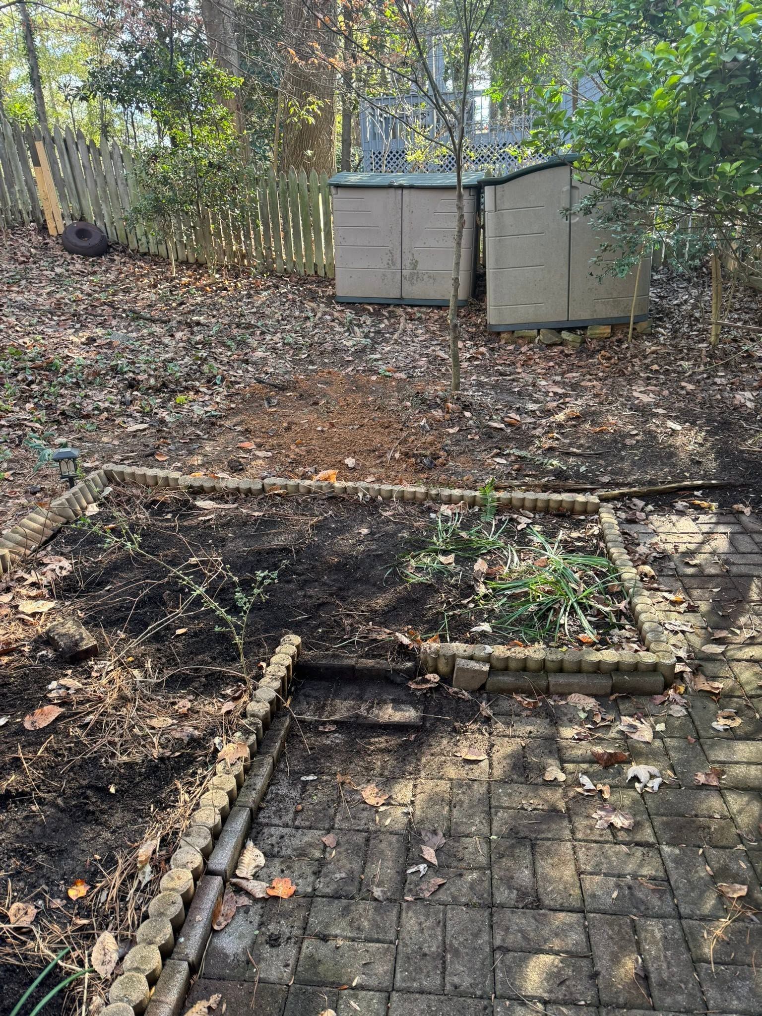 Backyard with fallen leaves, a dirt patch, paver edge, and two small storage sheds near trees