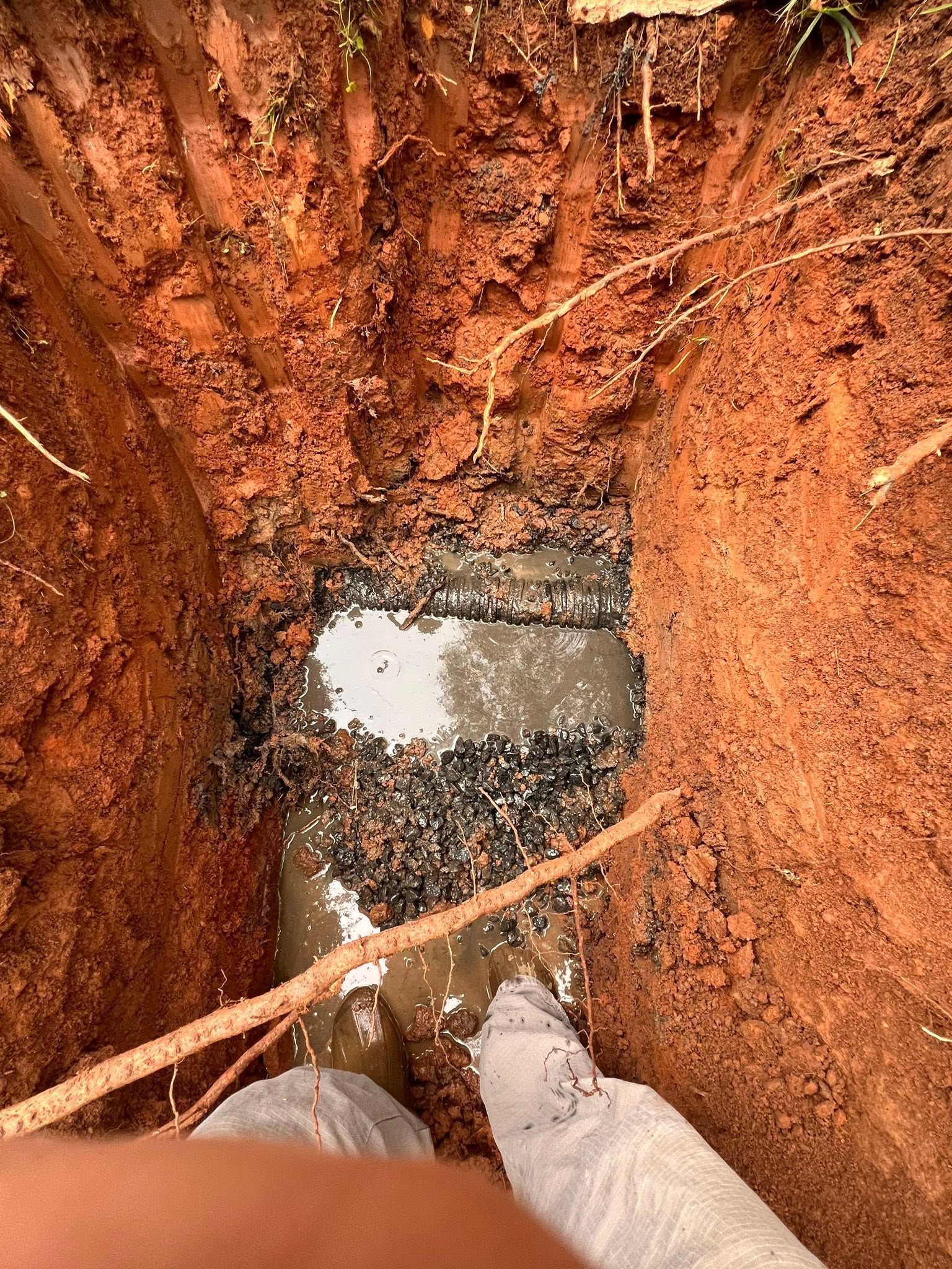 Looking down into a muddy trench with water at the bottom and a person’s boots visible
