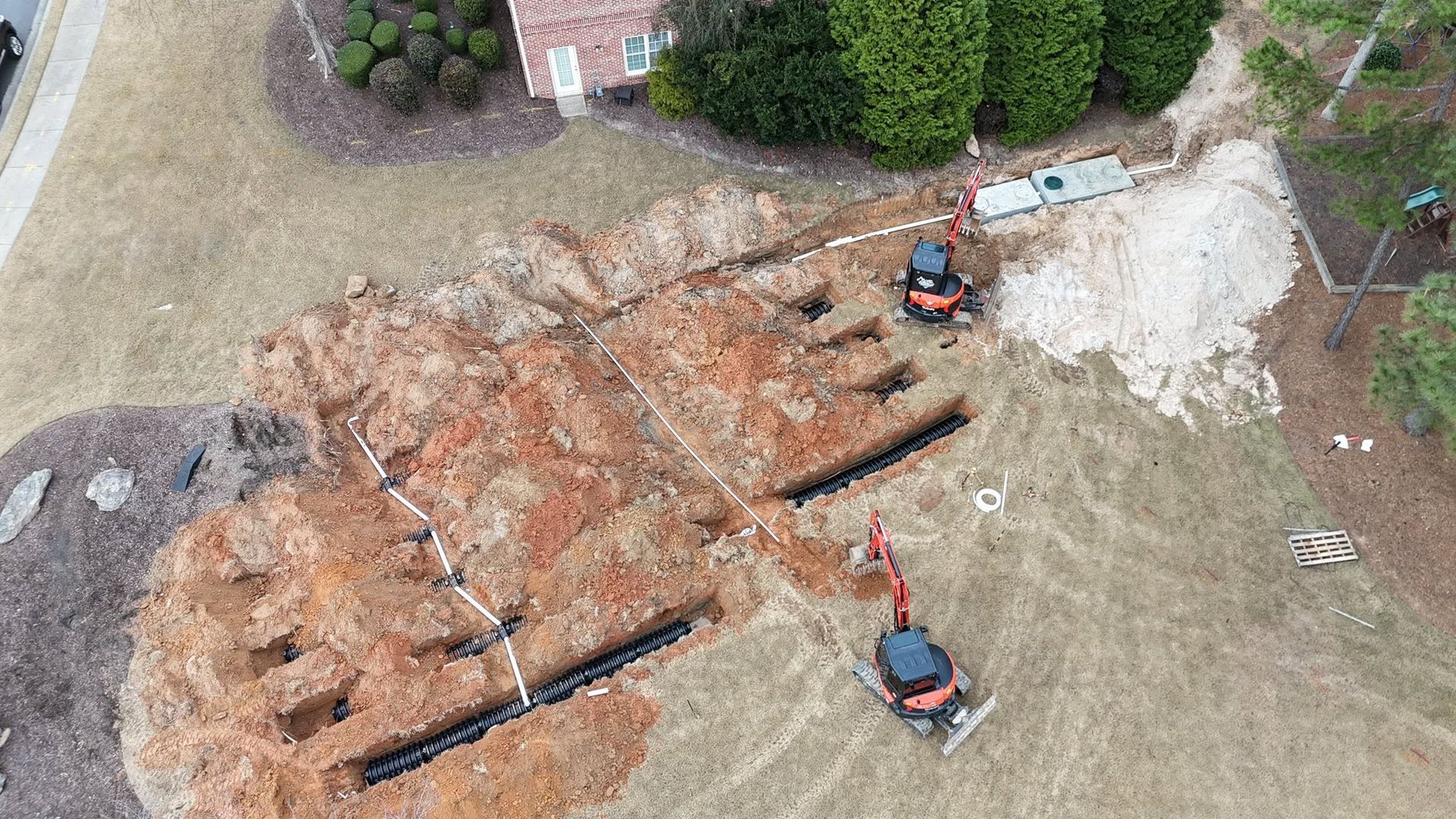 Aerial view of an excavated trench in a yard with two small construction machines and disturbed soil