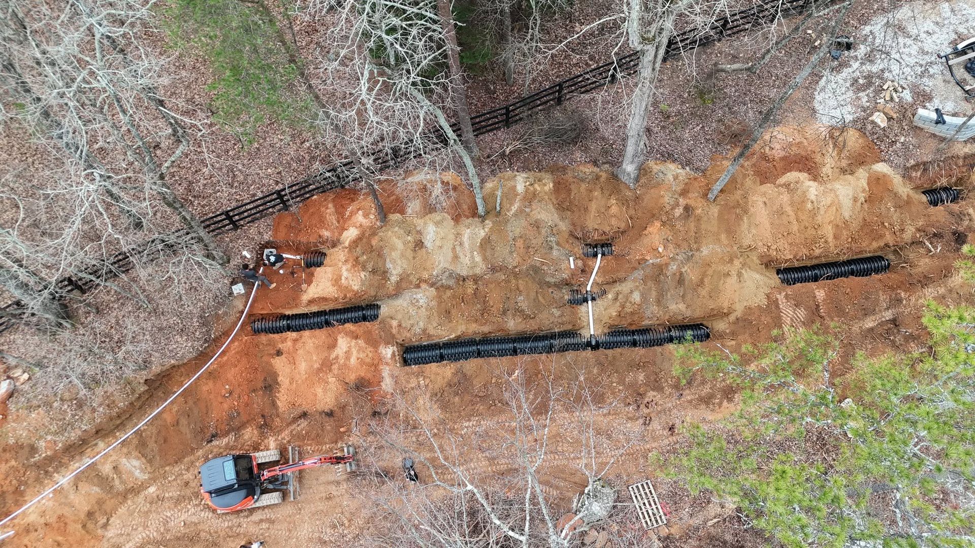 Aerial view of a muddy construction trench with black pipes and a red excavator beside leafless trees.