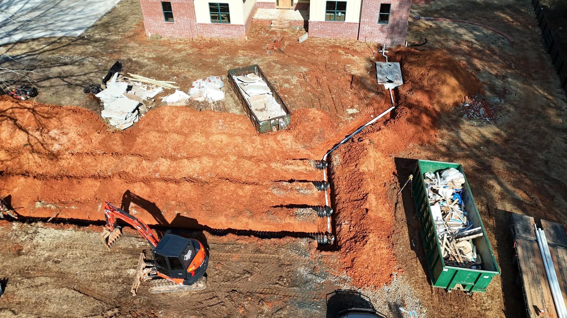 Excavator working on a dirt foundation beside a brick building, with a green dumpster nearby.