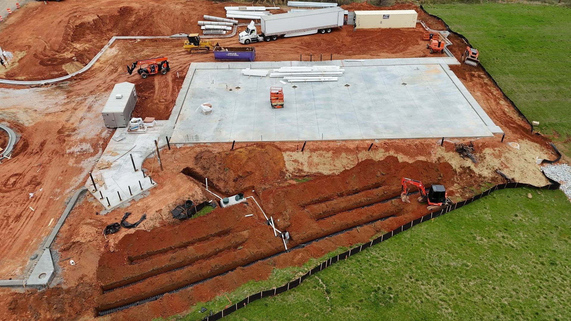Aerial view of a concrete foundation under construction on a red dirt site with earthmoving equipment.