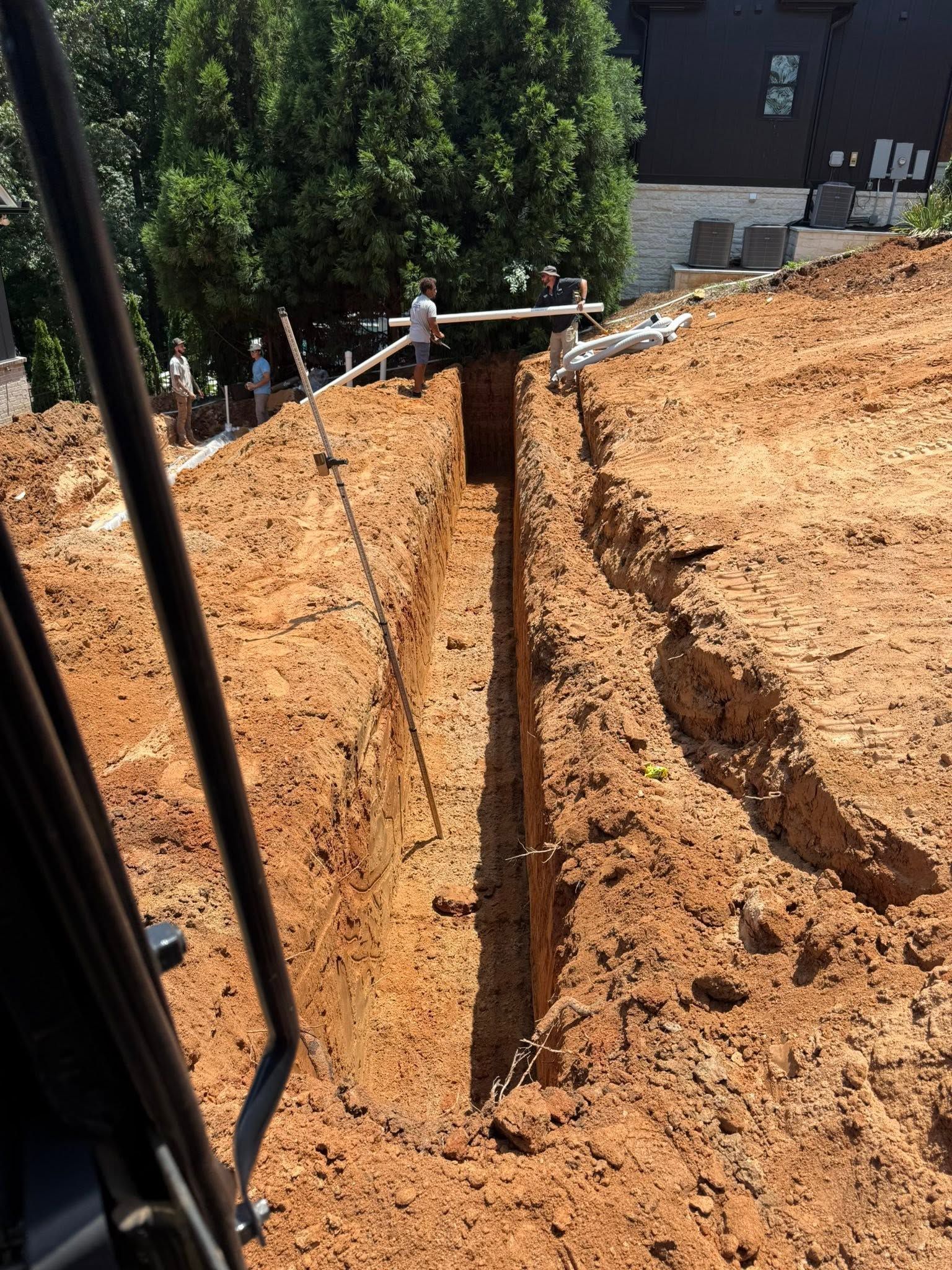 A deep trench in orange soil beside a building, with a vertical metal post and construction equipment nearby.